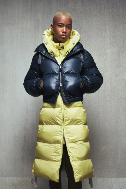 A woman with short hair poses in a two-tone navy and chartreuse puffer coat in front of a textured gray wall in a studio shot.