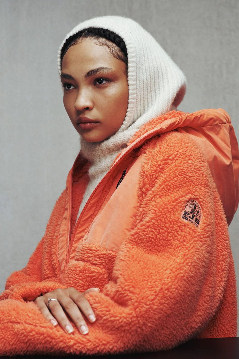 Woman poses in bright orange fleece jacket with knit hood and turtleneck against neutral backdrop, studio lighting, fashion portrait.