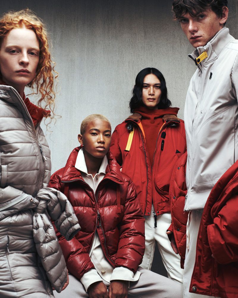 Group of four fashion models in winter puffer coats, two women and two men, in a studio setting with a concrete wall background.