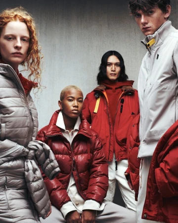 Group of four fashion models in winter puffer coats, two women and two men, in a studio setting with a concrete wall background.