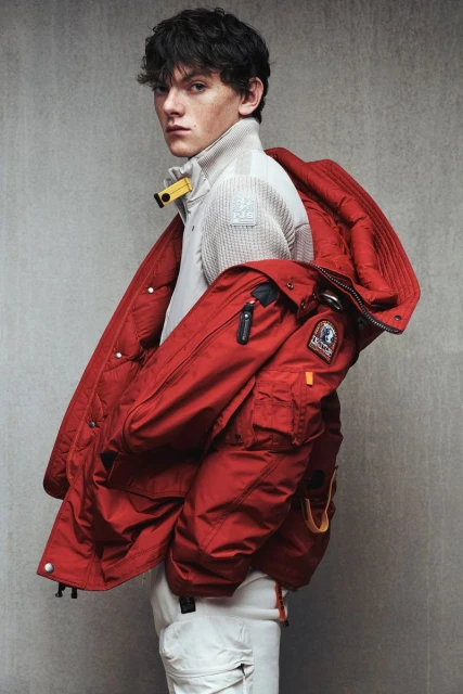 Young man posing indoors with a red winter jacket draped over his shoulders, wearing a beige turtleneck sweater and white pants.