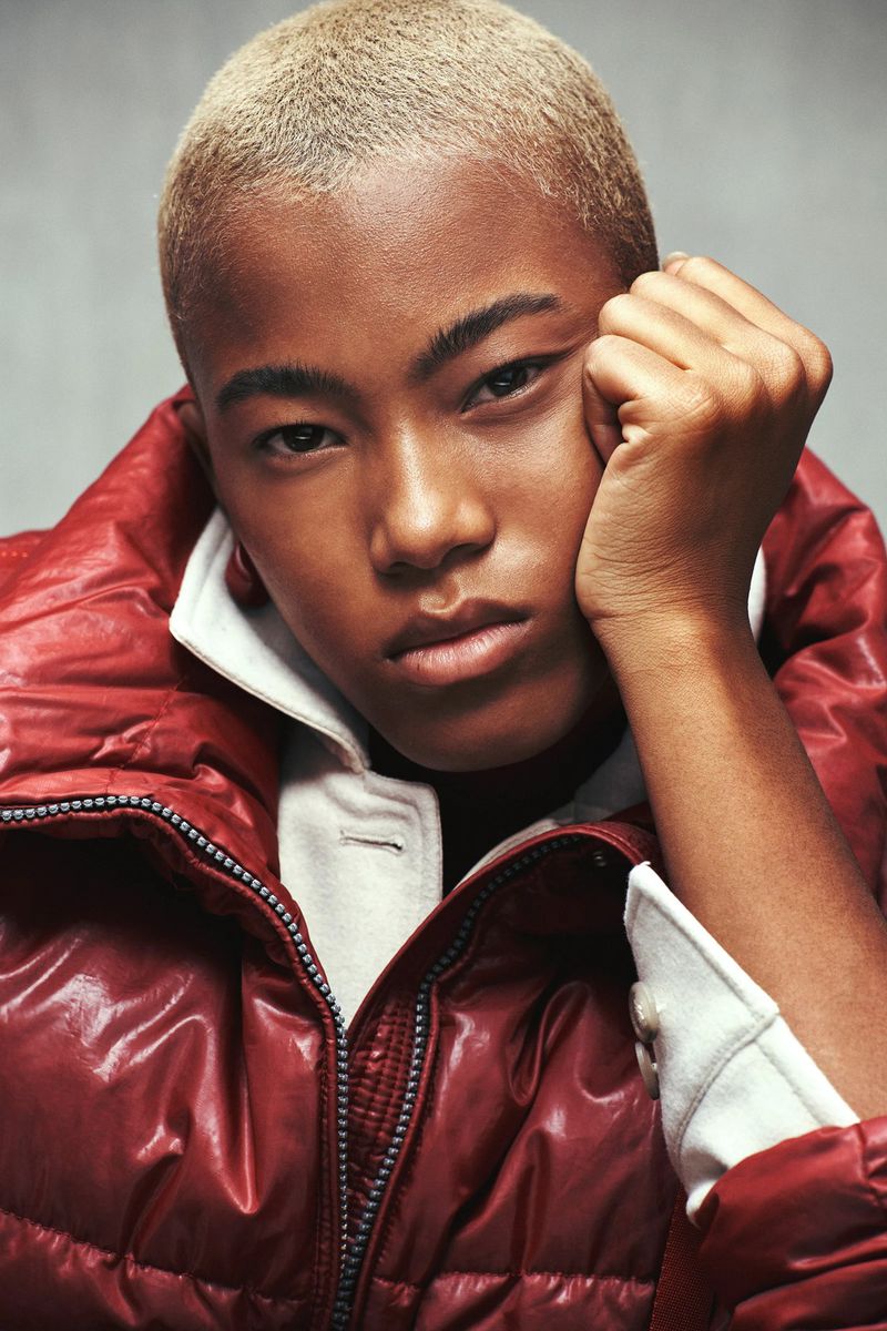 Close-up studio shot of a young adult posing in a red puffer jacket with a neutral expression and light blonde hair.