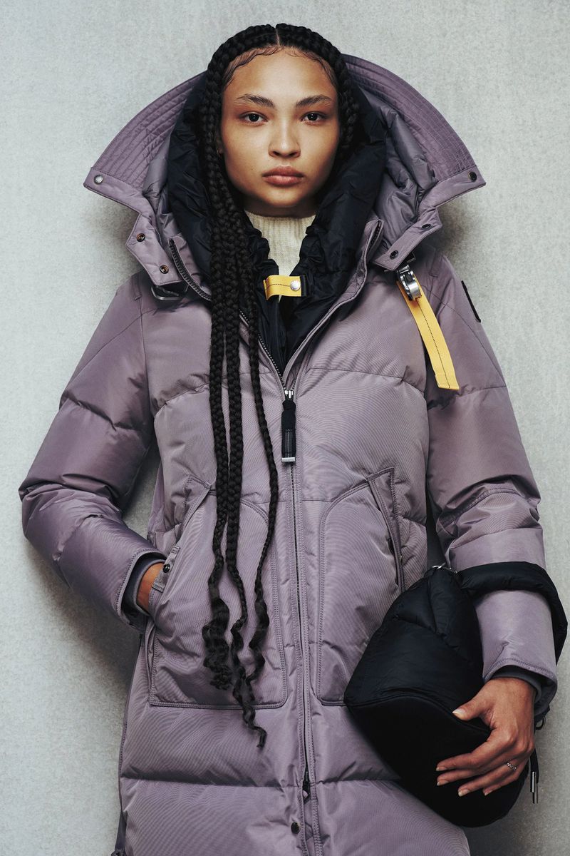 Fashionable young woman wearing a long lavender puffer coat with a hood and carrying a black handbag in a studio setting.
