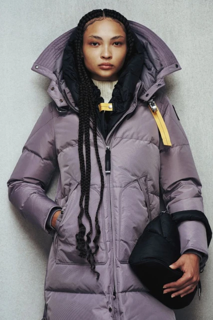 Fashionable young woman wearing a long lavender puffer coat with a hood and carrying a black handbag in a studio setting.