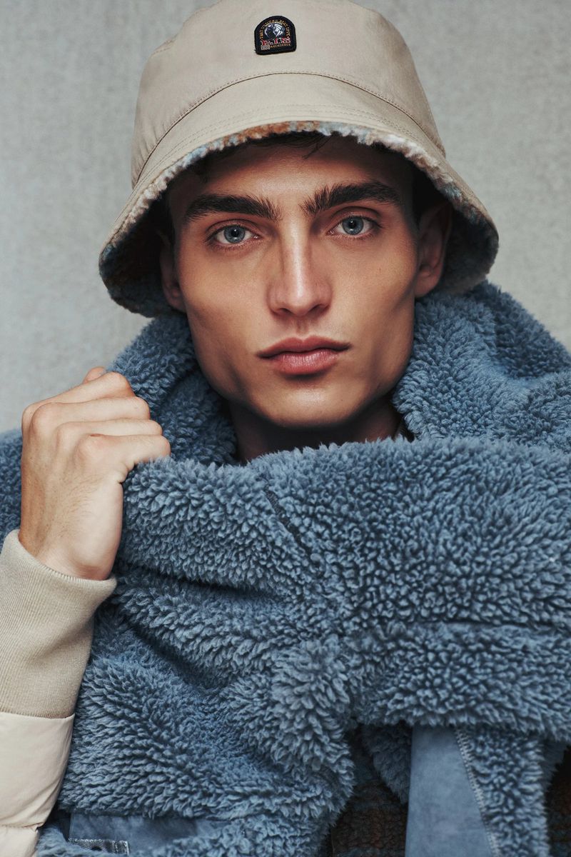 Portrait of a fair-skinned adult male wearing a beige bucket hat and a blue sherpa scarf in a close-up studio shot with a grey background.