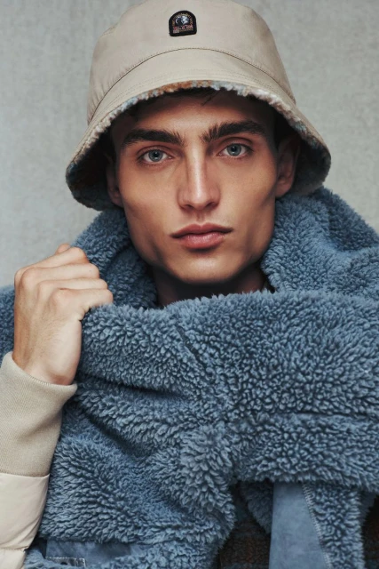 Portrait of a fair-skinned adult male wearing a beige bucket hat and a blue sherpa scarf in a close-up studio shot with a grey background.