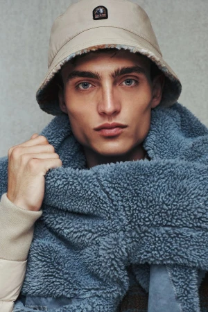 Close-up portrait of a male model wearing a beige bucket hat and blue fleece jacket against a neutral grey wall.