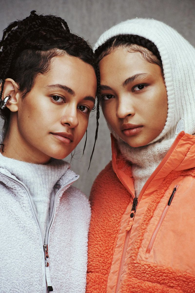 Close-up studio shot of two young women in fleece jackets, one in coral with a hood, the other in grey, both looking at the camera.