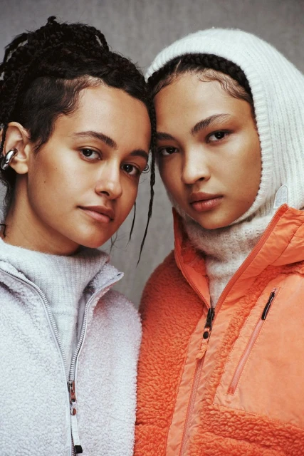 Close-up studio shot of two young women in fleece jackets, one in coral with a hood, the other in grey, both looking at the camera.