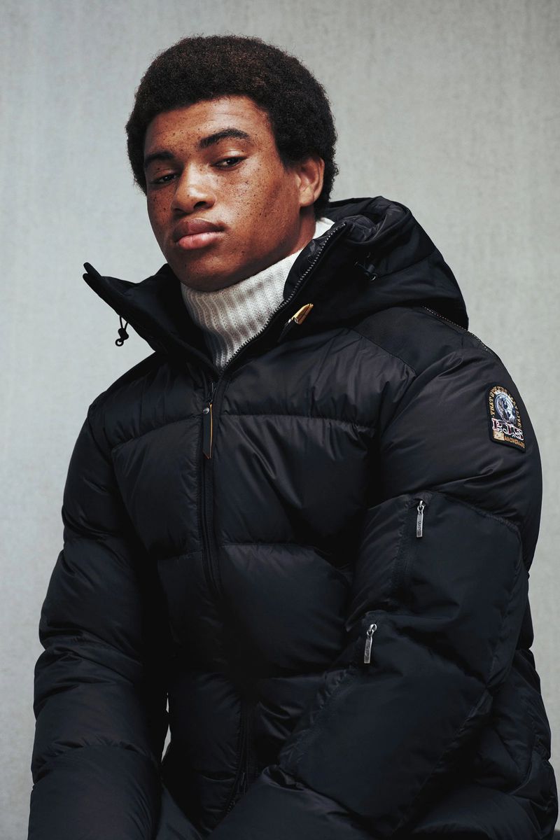 Man in black puffer jacket with fur-lined hood and white turtleneck sweater against a neutral background in medium studio shot.