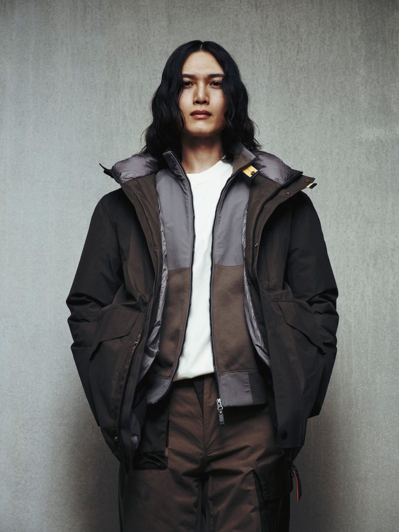A full-length portrait of a young Asian male wearing a down jacket and matching brown pants, posing in a minimalist indoor environment.