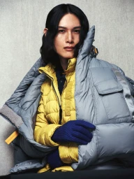 Model wearing a yellow puffer jacket under a large grey puffer coat and blue gloves against a textured background.
