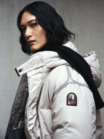 Model with dark wavy hair wearing an off-white puffer jacket and black scarf against a concrete texture wall.