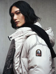 Model with dark wavy hair wearing an off-white puffer jacket and black scarf against a concrete texture wall.