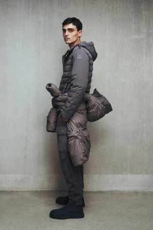 A man wearing a hooded grey puffer jacket with an additional padded coat tied around his waist against a concrete studio wall.