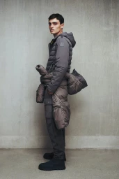 A man wearing a hooded grey puffer jacket with an additional padded coat tied around his waist against a concrete studio wall.