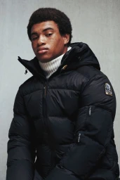 Black man wearing a black puffer jacket and white turtleneck against a plain textured background