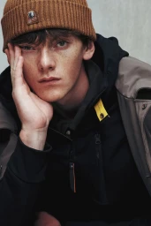 Close-up portrait of a male model wearing a brown beanie hat and dark jacket against a plain background