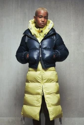 Model Pace Chen wearing a layered yellow long quilted coat and glossy navy cropped puffer jacket against a textured grey background.