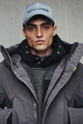 Model wearing a grey cap with branded text, a black hoodie, and a grey winter parka against a neutral concrete background