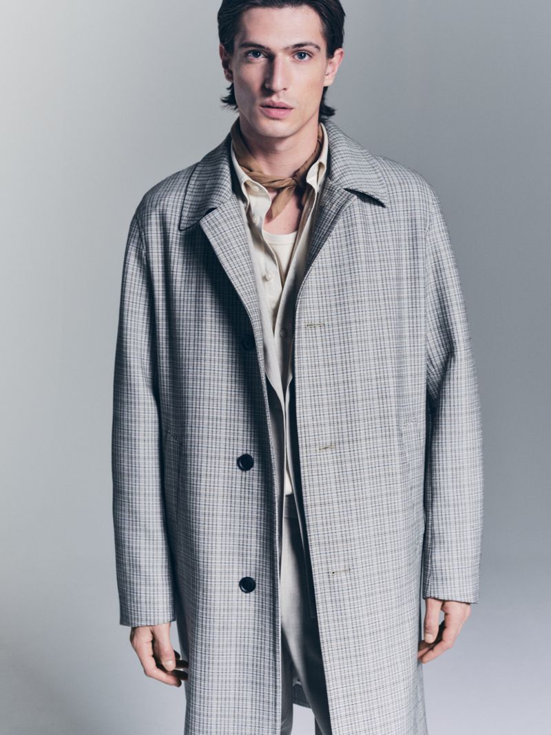 Man in beige shirt and patterned grey overcoat in a studio shot. He is gazing at the camera with a neutral expression and light skin.