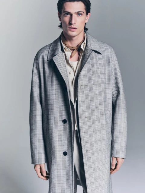 Man in beige shirt and patterned grey overcoat in a studio shot. He is gazing at the camera with a neutral expression and light skin.
