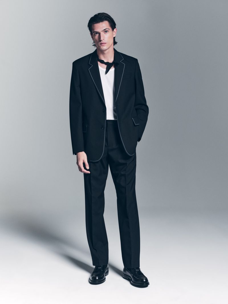 A handsome man in a black suit, crisp white t-shirt, and black kerchief poses in a studio, illuminated by bright, even lighting.