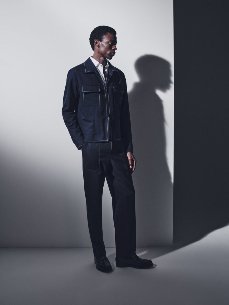 Dark-skinned adult male wearing a navy blue jacket and pants, stands in a photo studio with shadows on the wall, showcasing fashion.