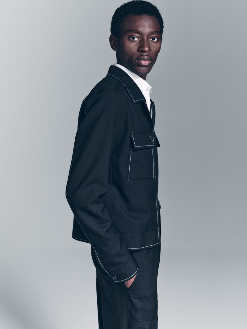 Portrait of a Black male fashion model in a dark jacket with white stitching and matching pants against a gray background in studio lighting.