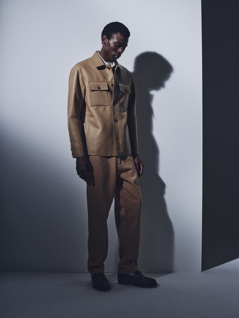 Model wearing a tan leather jacket and matching trousers standing against a light grey studio backdrop with a prominent shadow