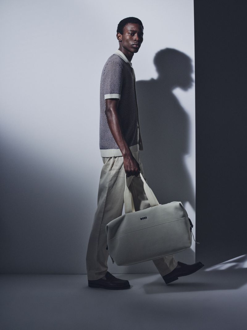 An adult male model walks in a grey, beige and white color graded photo studio wearing neutral-toned clothes while holding a light grey duffel bag.