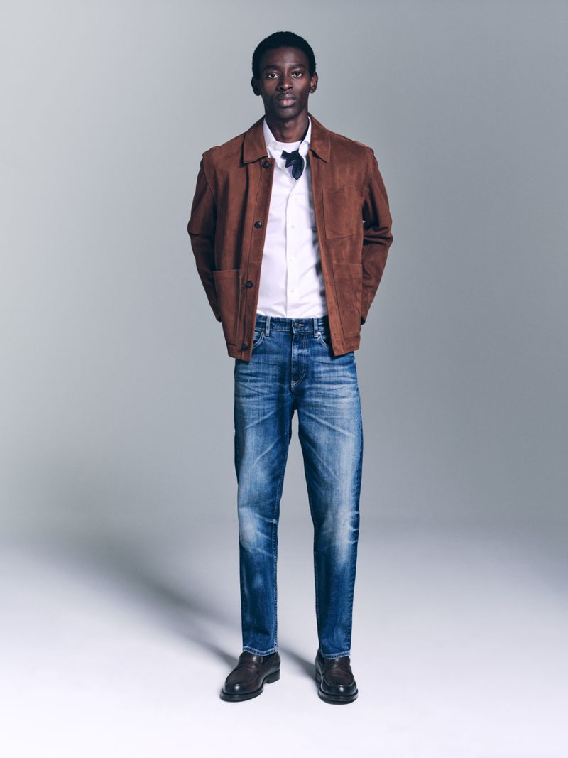 Full-length studio shot of an adult male fashion model in a brown jacket, white shirt, blue jeans, and black dress shoes against a grey backdrop.