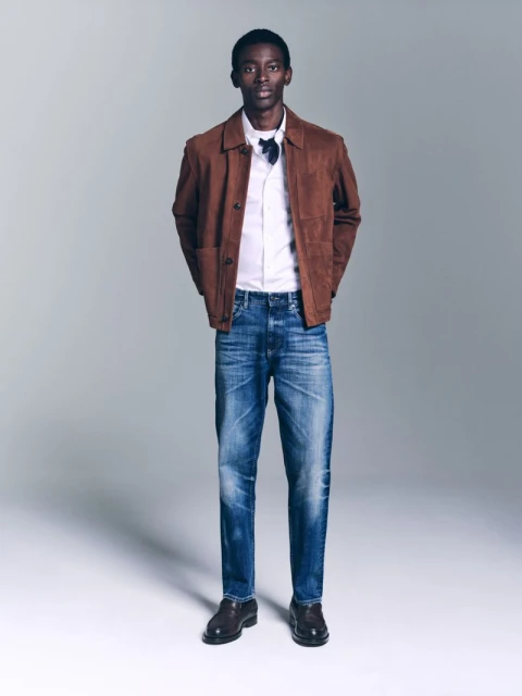Full-length studio shot of an adult male fashion model in a brown jacket, white shirt, blue jeans, and black dress shoes against a grey backdrop.