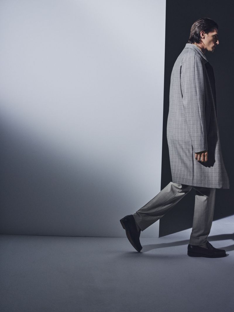A stylish adult man in a gray plaid overcoat is walking in a dimly lit, cool-toned indoor studio. The composition is minimalist and modern.
