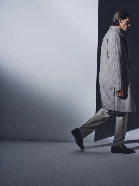 A stylish adult man in a gray plaid overcoat is walking in a dimly lit, cool-toned indoor studio. The composition is minimalist and modern.