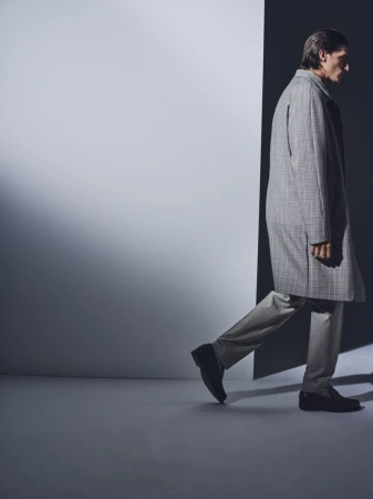 A stylish adult man in a gray plaid overcoat is walking in a dimly lit, cool-toned indoor studio. The composition is minimalist and modern.