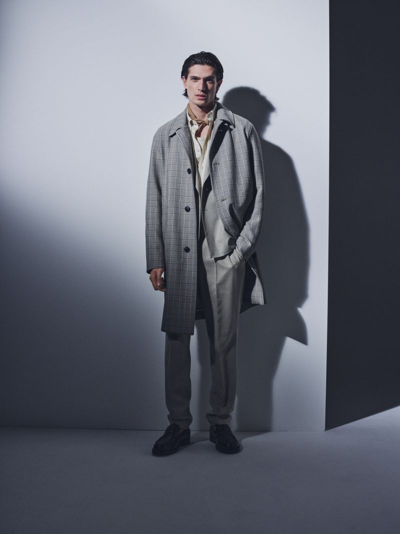 Full-length portrait of an adult man in a grey overcoat, light suit, and dark shoes standing against a gradient grey background in a studio.
