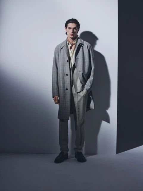 Full-length portrait of an adult man in a grey overcoat, light suit, and dark shoes standing against a gradient grey background in a studio.