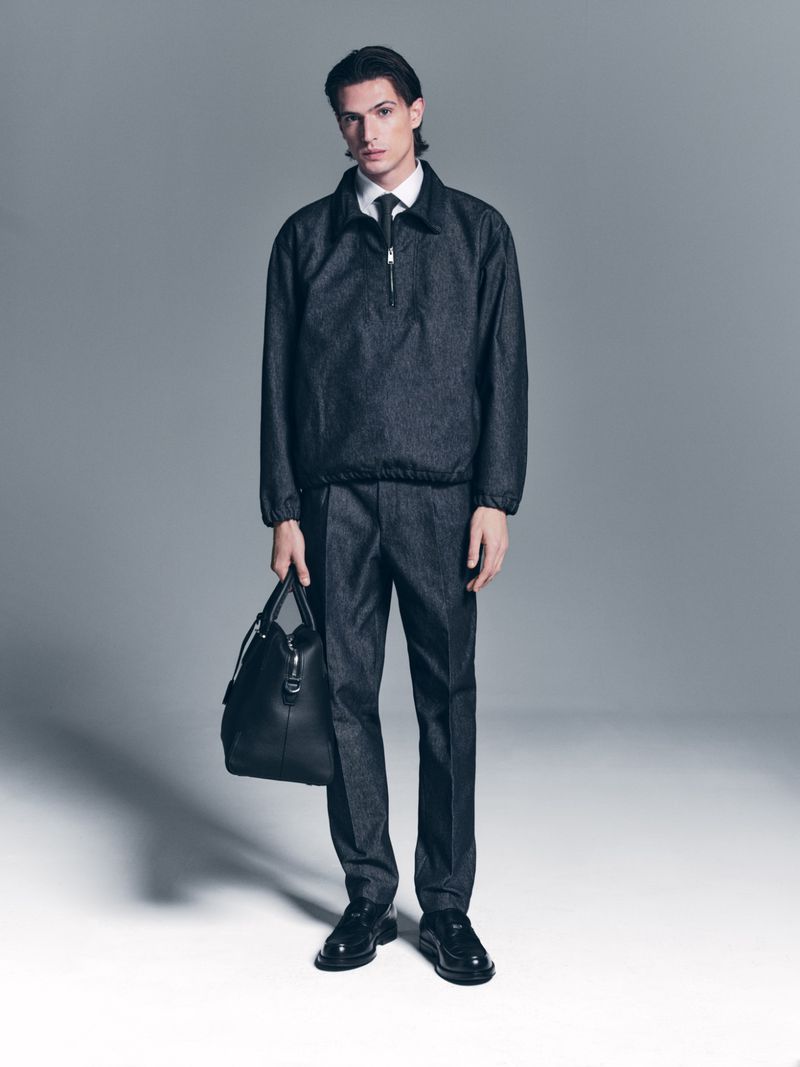 Full shot of a stylish adult male model in a gray outfit and black leather bag, captured in a studio setting.