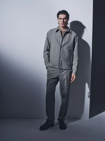 Model Edoardo Sebastianelli posing in a grey checked jacket and matching trousers against a studio wall with a sharp shadow.