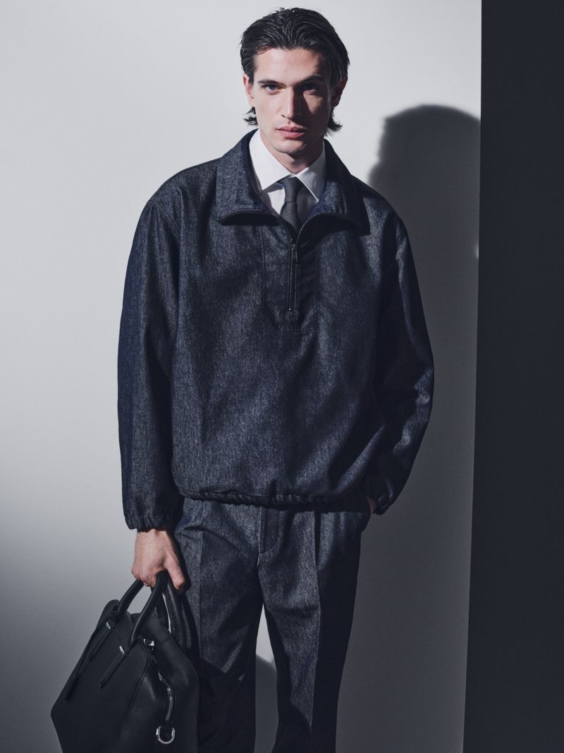 Adult male poses in a light, neutral indoor setting wearing a denim jacket and pants, holding a black leather bag. Formal attire.