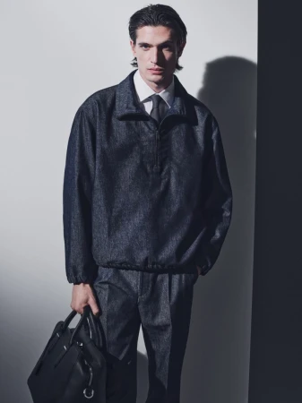 Adult male poses in a light, neutral indoor setting wearing a denim jacket and pants, holding a black leather bag. Formal attire.