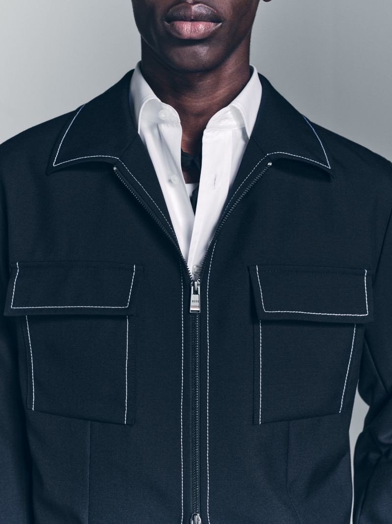 A close-up shot of a black male wearing a white dress shirt underneath a black jacket with white stitching detail.