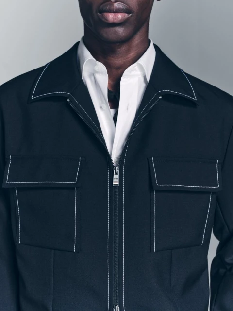 A close-up shot of a black male wearing a white dress shirt underneath a black jacket with white stitching detail.