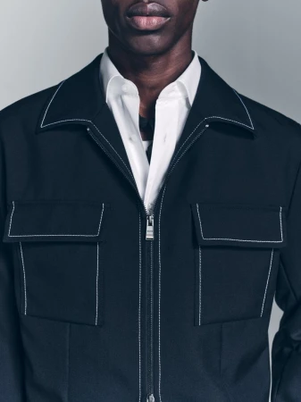 A close-up shot of a black male wearing a white dress shirt underneath a black jacket with white stitching detail.