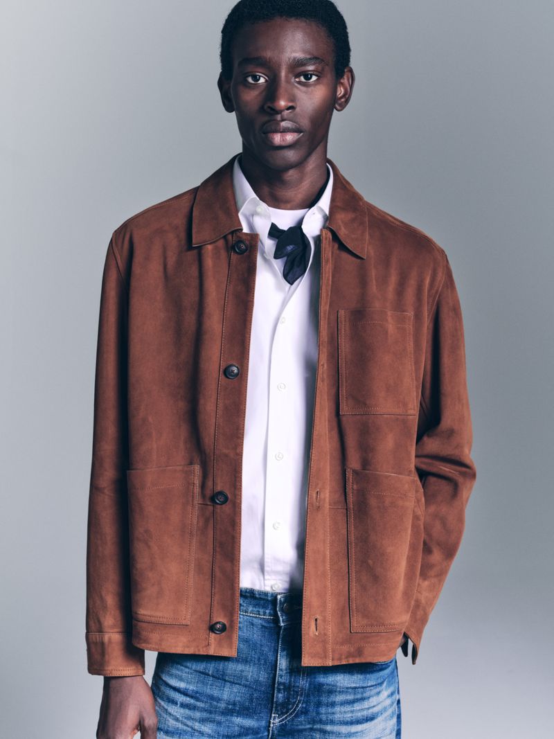 Adult male in a fashion shot wearing a brown suede jacket, white shirt, and blue jeans against a neutral backdrop in soft studio lighting.