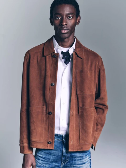 Adult male in a fashion shot wearing a brown suede jacket, white shirt, and blue jeans against a neutral backdrop in soft studio lighting.