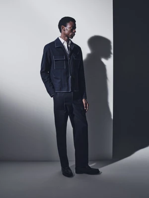 Model wearing a dark utility jacket and trousers standing against a pale studio wall with a sharp shadow.