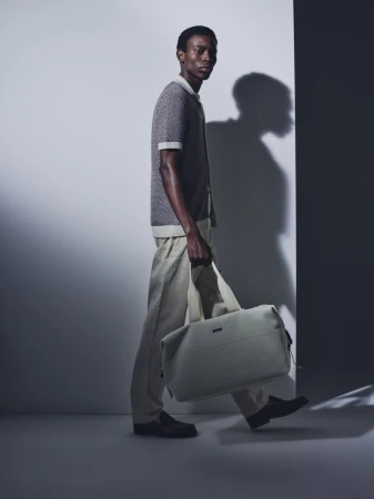 Model Adamu Bulus walking with a cream duffle bag in a knit polo and beige trousers against a studio wall.