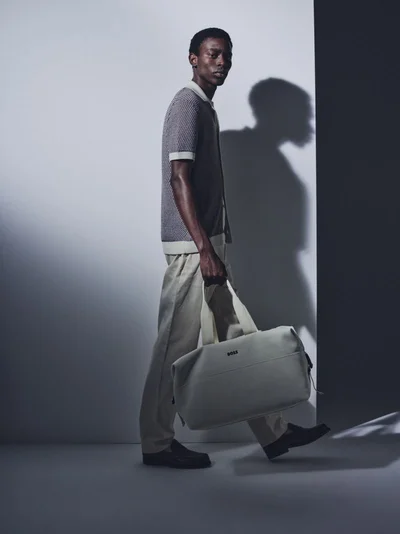 Model Adamu Bulus walking with a cream duffle bag in a knit polo and beige trousers against a studio wall.